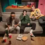 Person sitting on couch in dim room illuminated by TV screen, surrounded by snacks, late at night