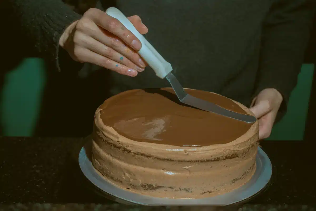 Hands spreading chocolate ganache over layers of assembled cheesecake on a serving board