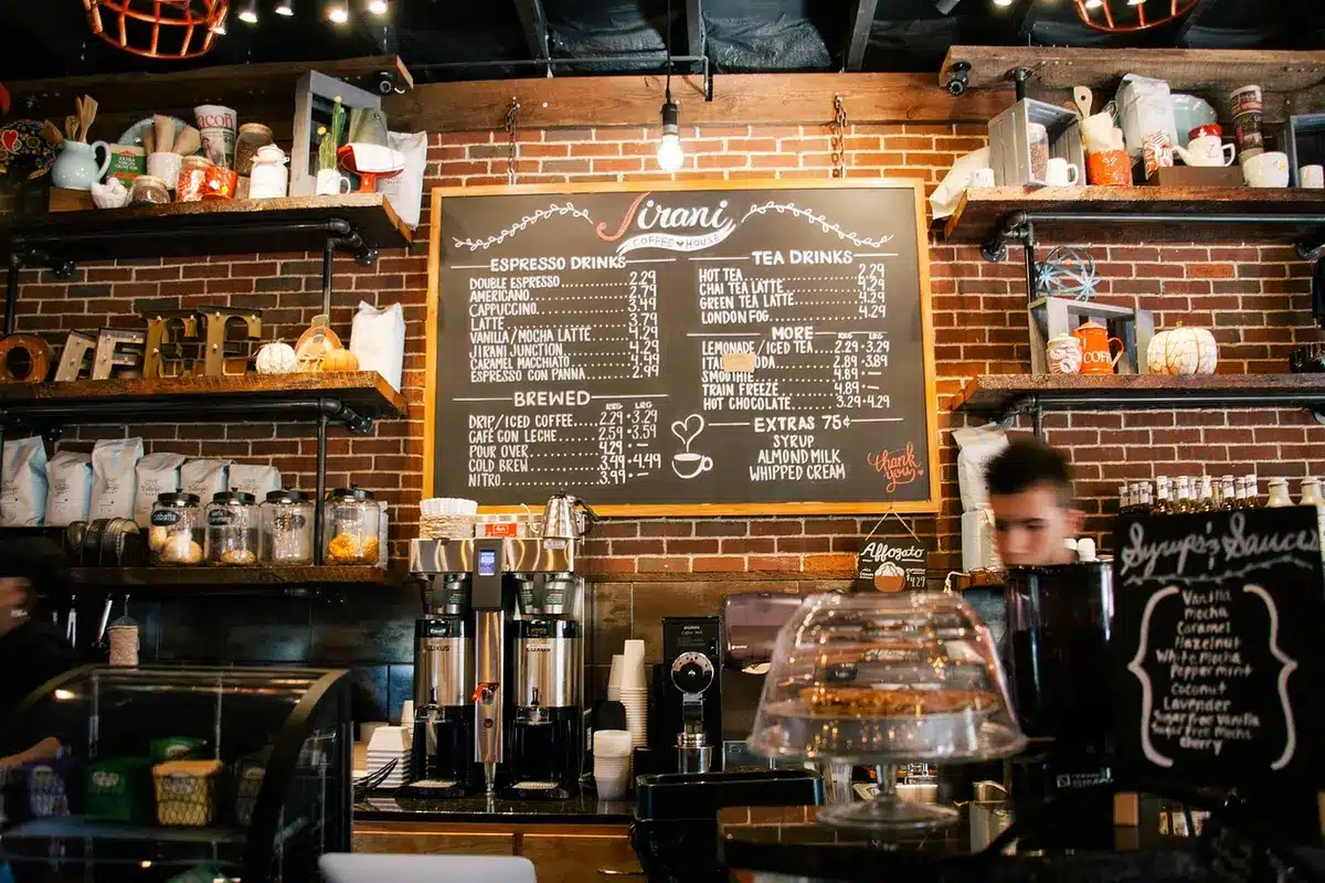 Barista steaming milk at a sunlit counter with a latte and syrup bottle